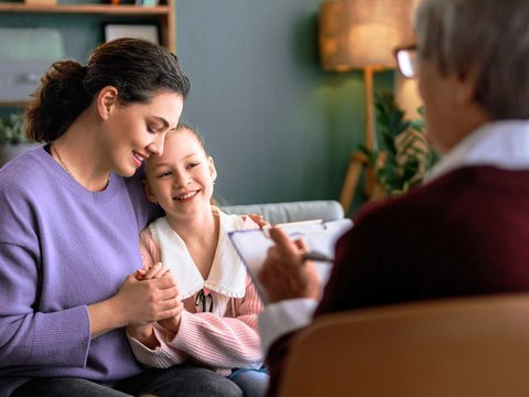 Eine Frau und ein Mädchen sitzen auf einem Sofa. Die Frau hält das Mädchen im Arm. Beide lächeln Ihnen gegenüber sitzt eine Person und notiert etwas auf einem Klemmbrett.