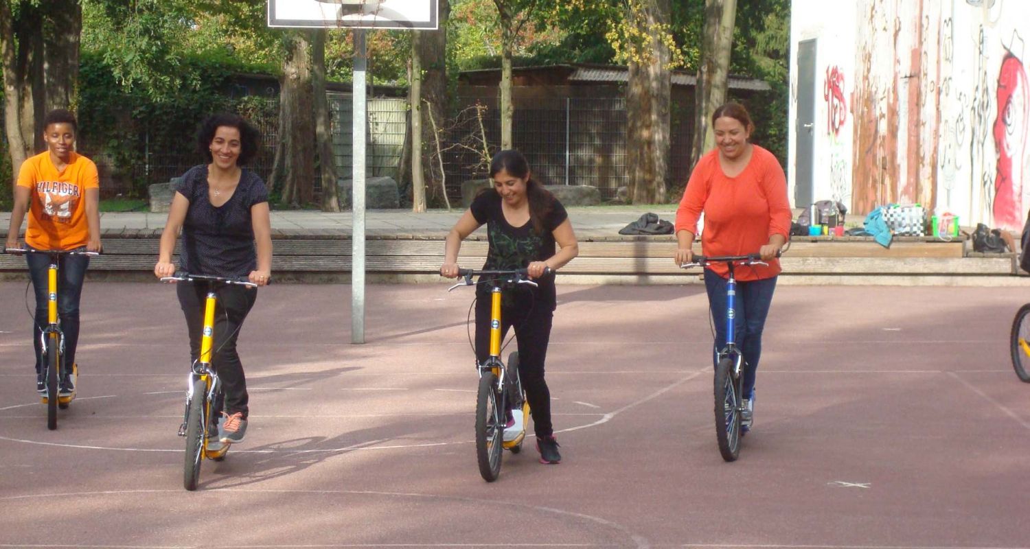 Vier Frauen üben auf einem Basketballplatz auf Rollern zu fahren.