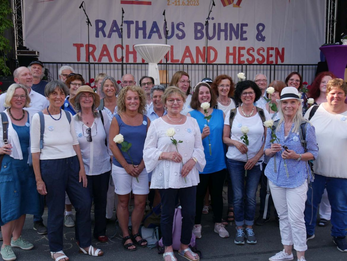 Der Chor All inklusiv steht vor der Rathaus Bühne auf dem Hessentag in Pfungstadt. Die Männer und Frauen lächeln in die Kamera. Einige haben weiße Rosen in der Hand.