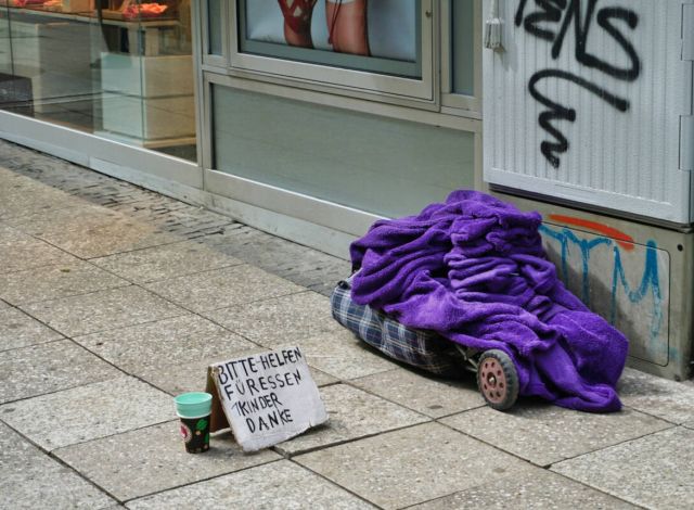 Decke und Tasche eines Obdachlosen liegen auf der Straße, mit Schild "Bitte helfen" und Becker für Geldspenden