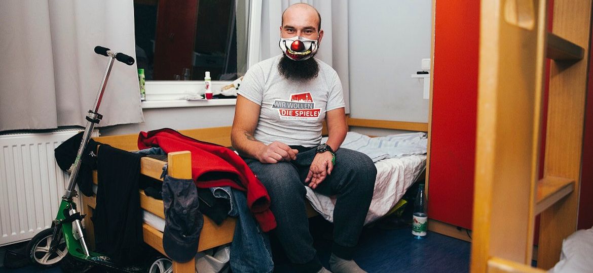 Blick in ein Zimmer der Wohnungslosenhilfe mit Mann mit Maske auf einem Bett sitzend